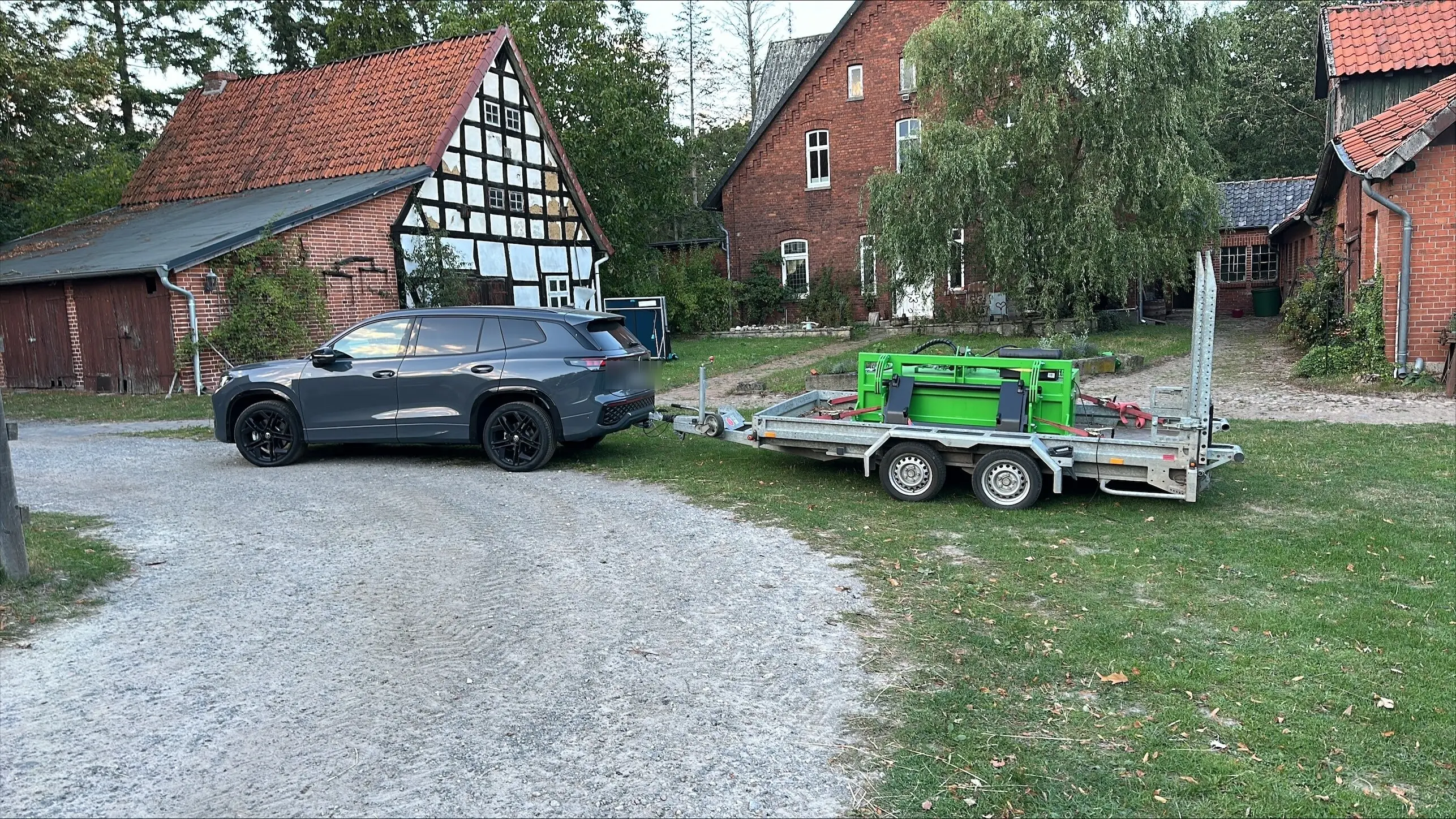 Zugfahrzeug mit Anhänger und grünem FARM-SERVANT ECO Abmistgerät auf dem Hofplatz vor einem Fachwerkhaus